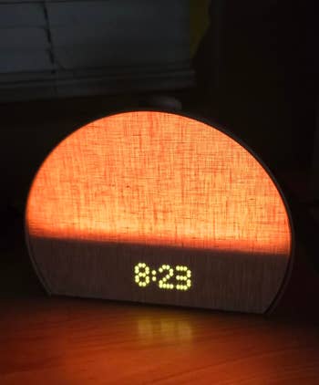 Digital clock on a bedside table displaying 8:23, illuminated with a warm glow from a fabric-covered light