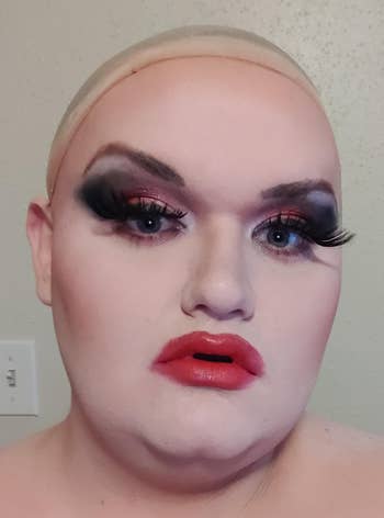 Person with dramatic eye makeup and bold lips poses with hand on chest, showcasing beauty and self-expression style for a shopping article