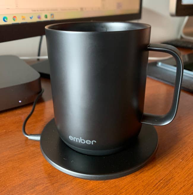 A black Ember mug on a charging coaster sits on a wooden desk in front of a computer monitor