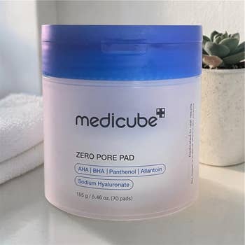 Medicube Zero Pore Pad container displayed on a bathroom counter with a towel and plant in the background