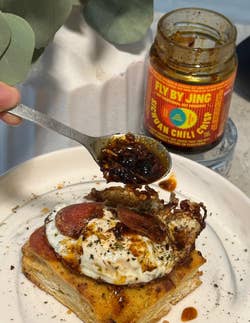 A spoonful of chili crisp being placed on a fried egg with pepperoni atop a slice of bread, next to a jar labeled