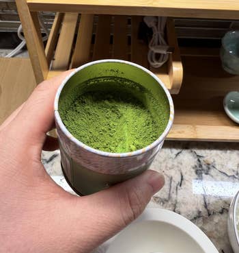 Hand holding a container of green tea powder on a kitchen counter, with shelves and a mug in the background