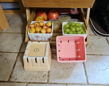 Wooden fruit stand with various fruits, including cherries and grapes, on a tiled countertop. Two empty crates are in the foreground