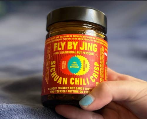 A hand holds a jar of Fly By Jing Sichuan Chili Crisp, a spicy condiment, against a soft blue background