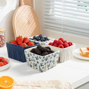 Three ceramic berry baskets with strawberries, blueberries, raspberries, and blackberries on a kitchen counter near sliced bread and an orange