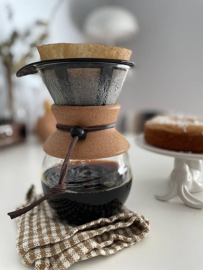 Glass pour-over coffee maker with a leather collar, sitting on a checked cloth. A cake on a stand is blurred in the background