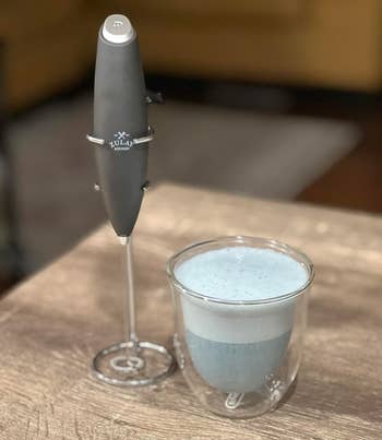 Milk frother with stand on a table beside a glass cup filled with frothy beverage, suggesting a home coffee experience