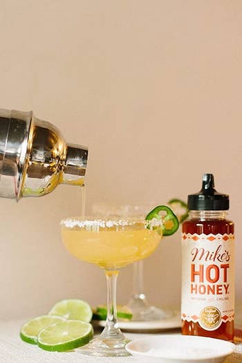Someone pouring a cocktail out of a shaker into a glass with Mike's Hot Honey next to the glass