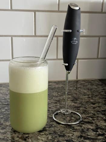 Glass of frothy matcha latte beside a milk frother on a kitchen counter
