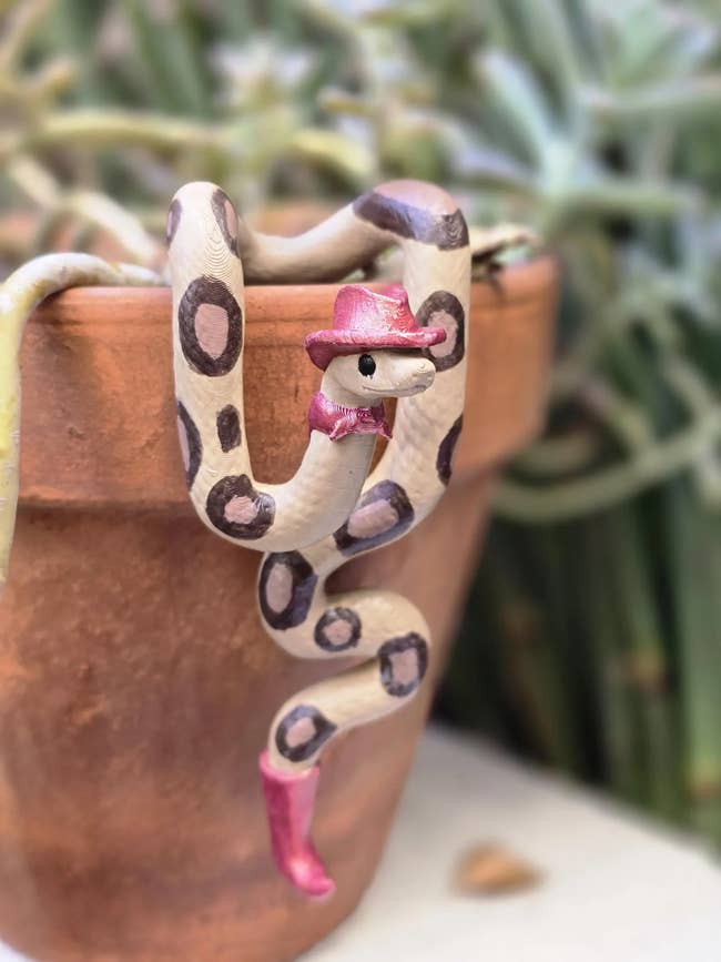 Decorative sculpture of a snake with circular spots, wearing a pink hat and boot, draped over a plant pot