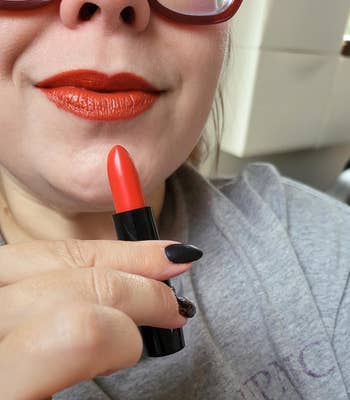 Person wearing sunglasses applies red lipstick, holding it near their lips. They wear a casual top