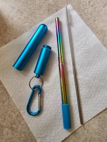 Collapsible reusable straw with a rainbow finish and blue case on a napkin; includes a cleaning brush and carabiner for portability