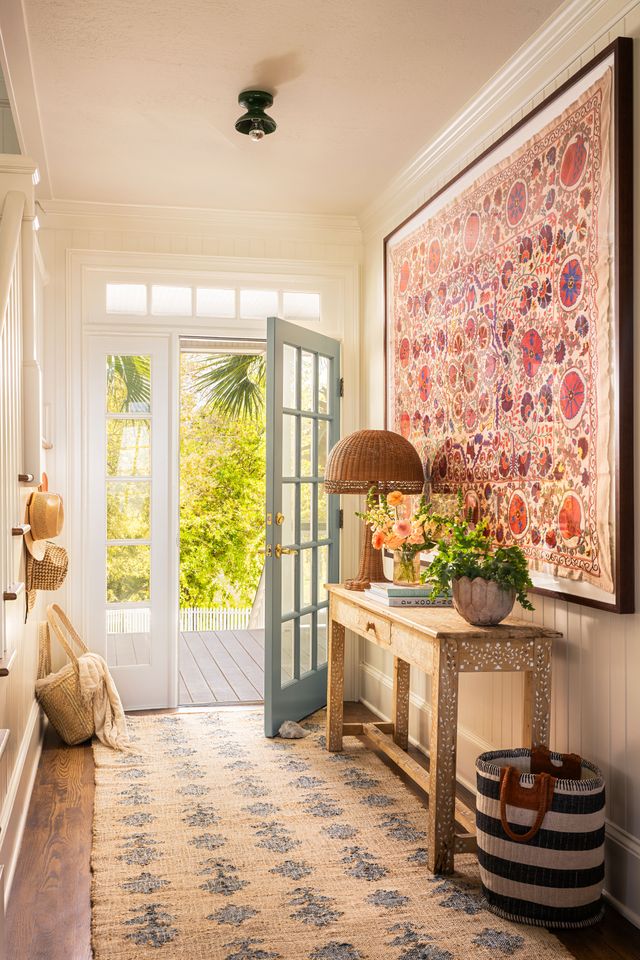 A brightly lit entryway with a large rug ornate wall art wooden table and glass door leading to an outdoor area filled with greenery