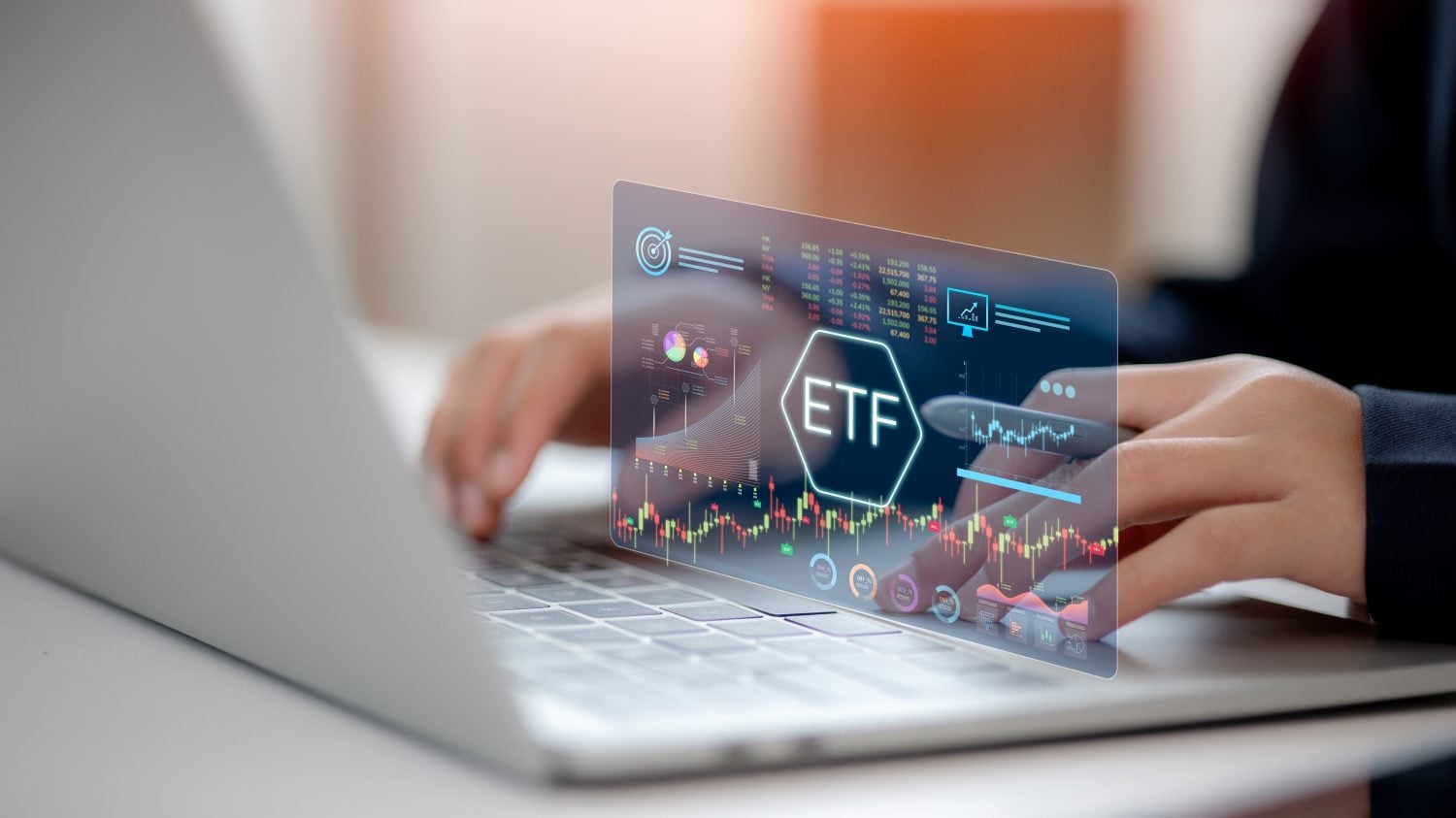 A person's hands are seen typing on a silver laptop. Floating above the keyboard is a transparent, blue-tinted holographic display showing financial information, including candlestick charts, pie graphs, numerical data, and the letters 'ETF' centered in a glowing hexagon.