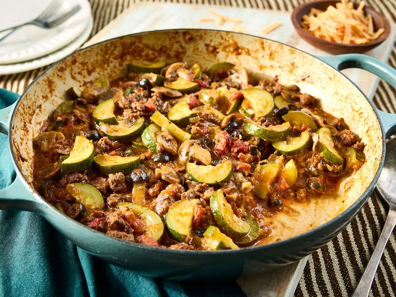 A skillet containing a zucchini and ground beef dish with visible tomatoes and spices placed on a table with a striped background