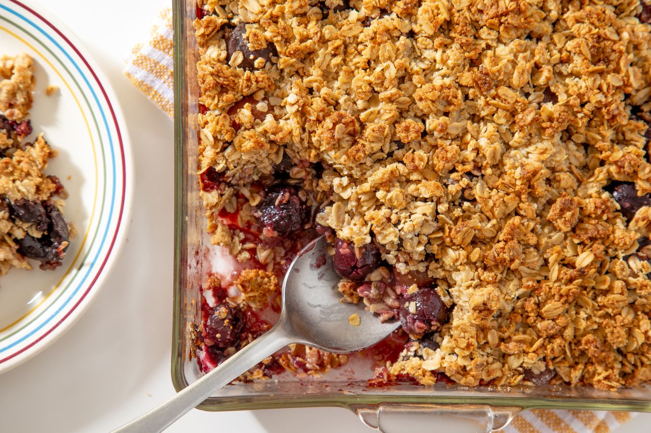 A dish of cherry crisp with a serving spoon and a portion served on a striped plate nearby