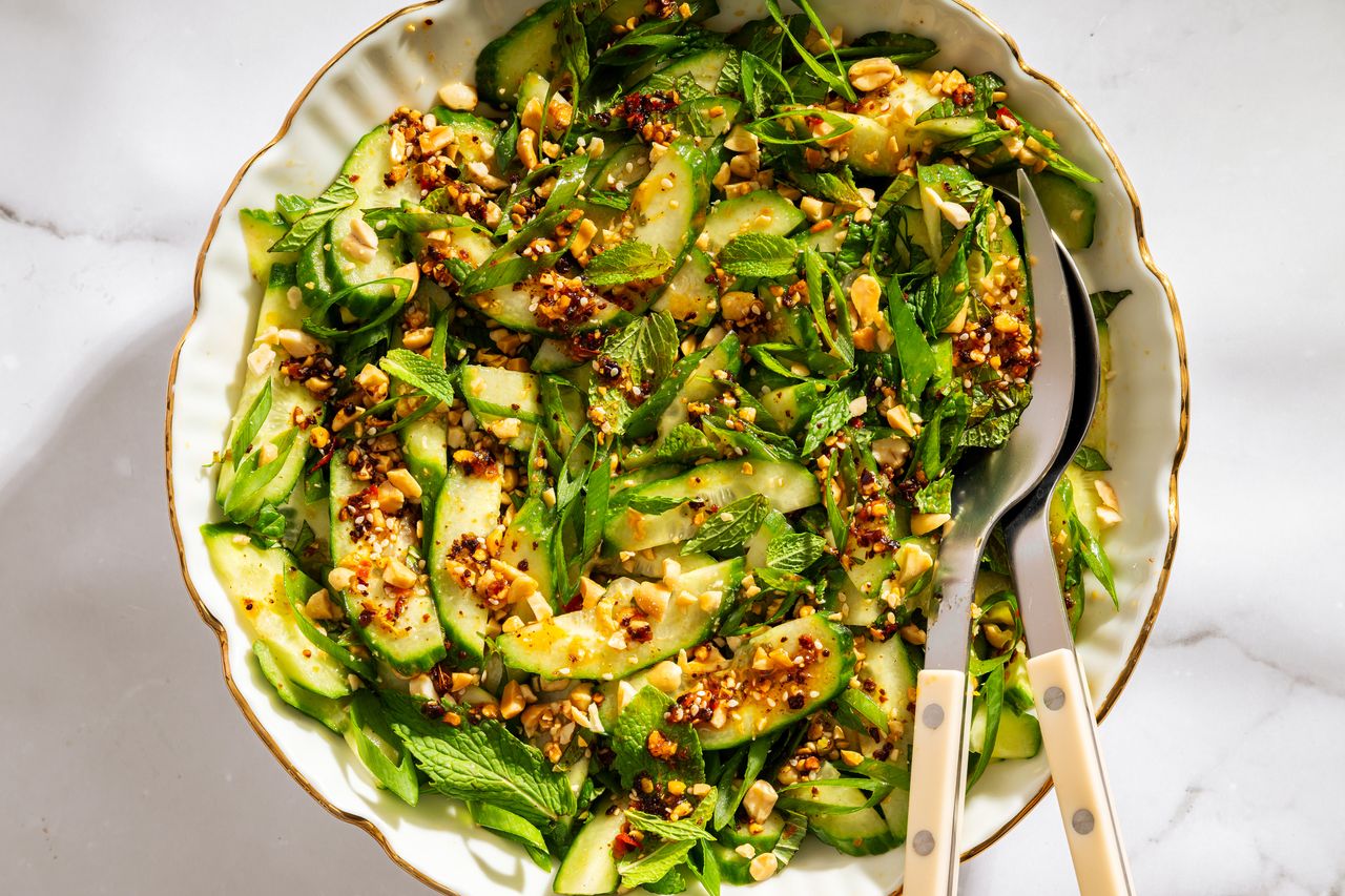 A bowl of spicy cucumber salad garnished with herbs and seeds with serving utensils