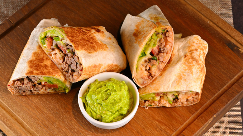 Halved burritos on wooden board with small bowl of guacamole