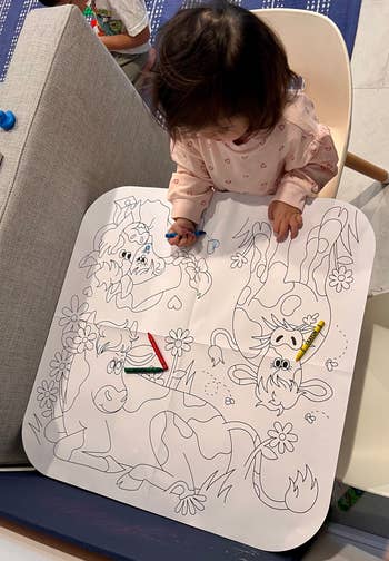 Child coloring a large illustrated mat featuring cartoon cows and flowers, with scattered crayons