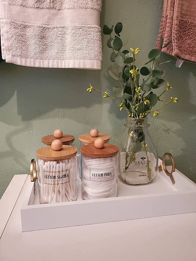 a reviewer photo of the labeled glass jars with wood lids filled with q-tips and cotton pads on a tray with a vase of eucalyptus