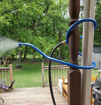 A garden hose tied to a porch post, spraying mist over a backyard with trees, a swing, and a wooden deck