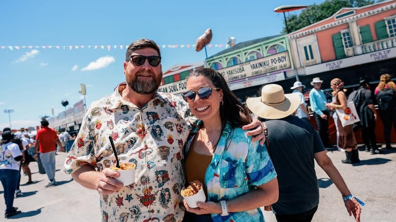 A couple posing to the camera while holding food with a variety of concession stands int he background