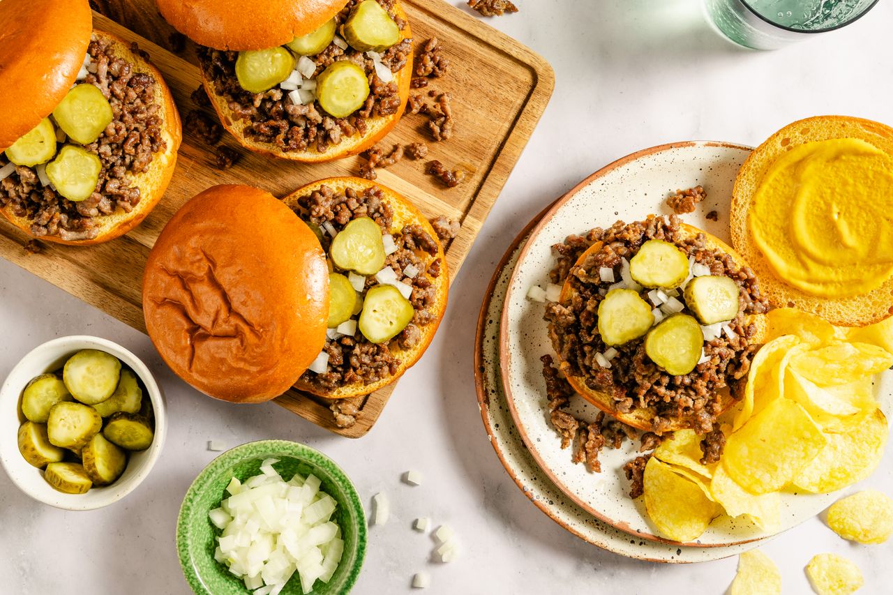 Loose meat sandwiches with pickles and onions on buns served with chips and mustard on a table