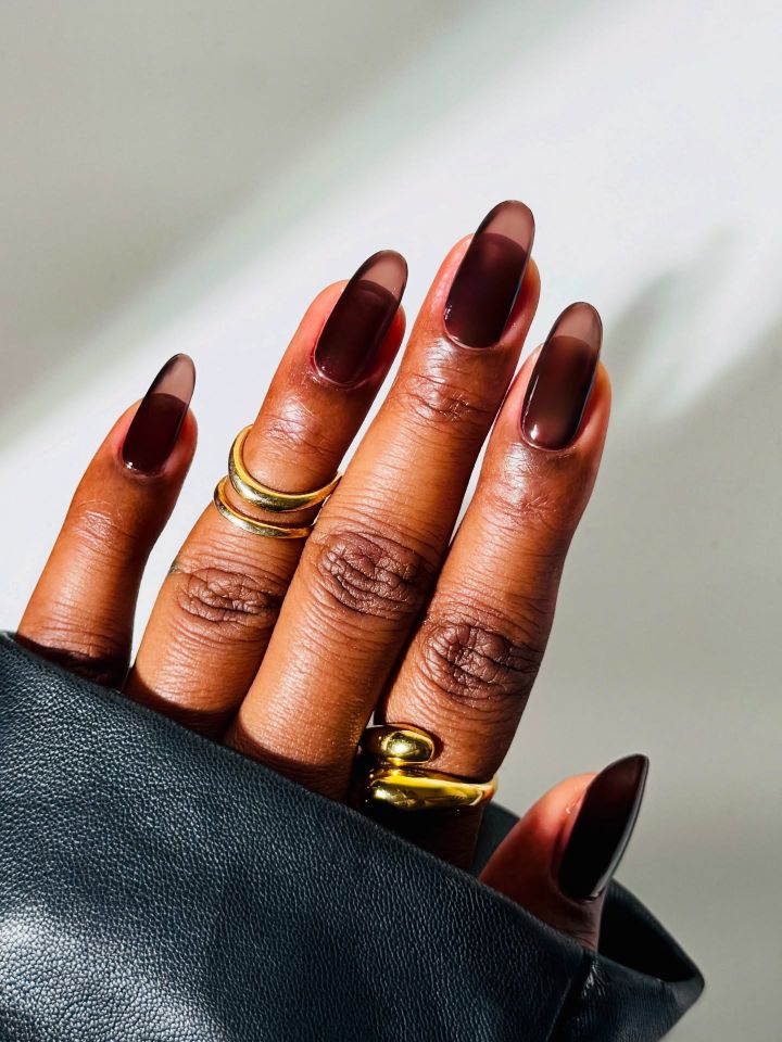 A hand with manicured nails featuring long dark transparent polish adorned with multiple gold rings wearing a black leather sleeve