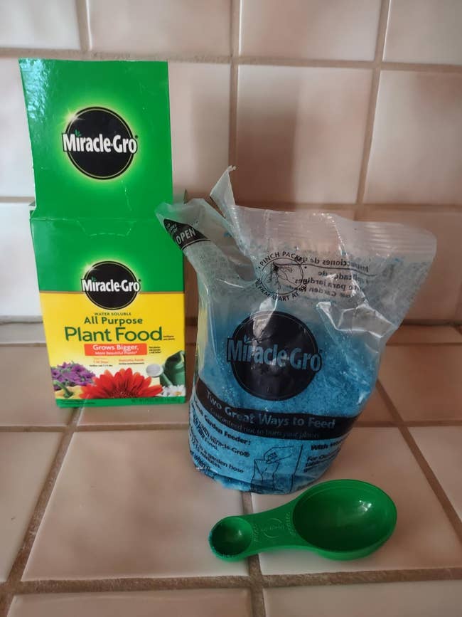 Miracle-Gro All Purpose Plant Food box, blue granules in a plastic bag, and a green scoop on tile countertop