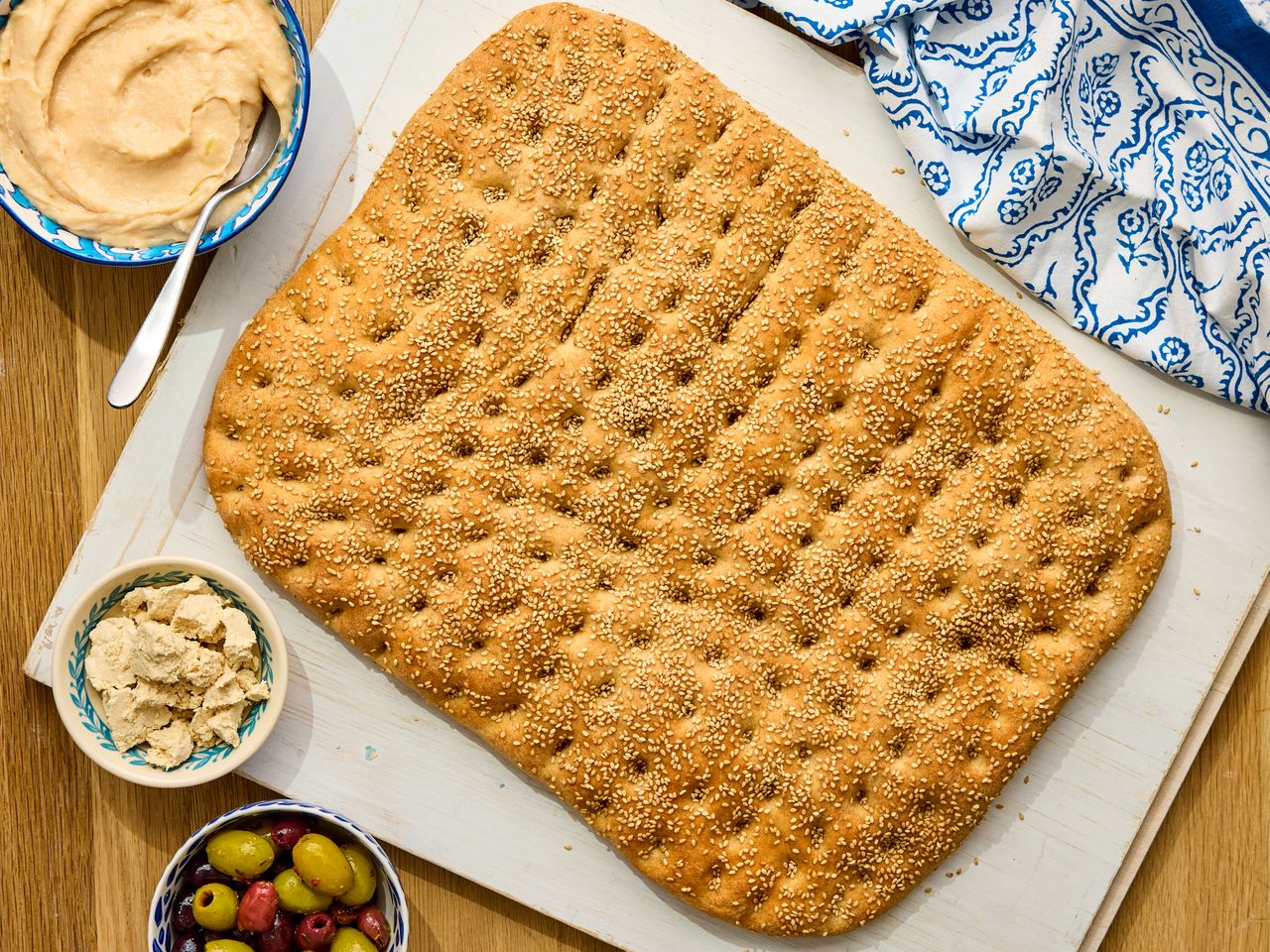 A sheet of freshly baked focaccia on a surface surrounded by bowls of hummus and olives