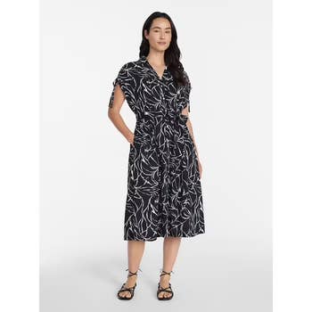 Person wearing a stylish knee-length dress with a bold leaf pattern, standing with hands in pockets, paired with strappy sandals