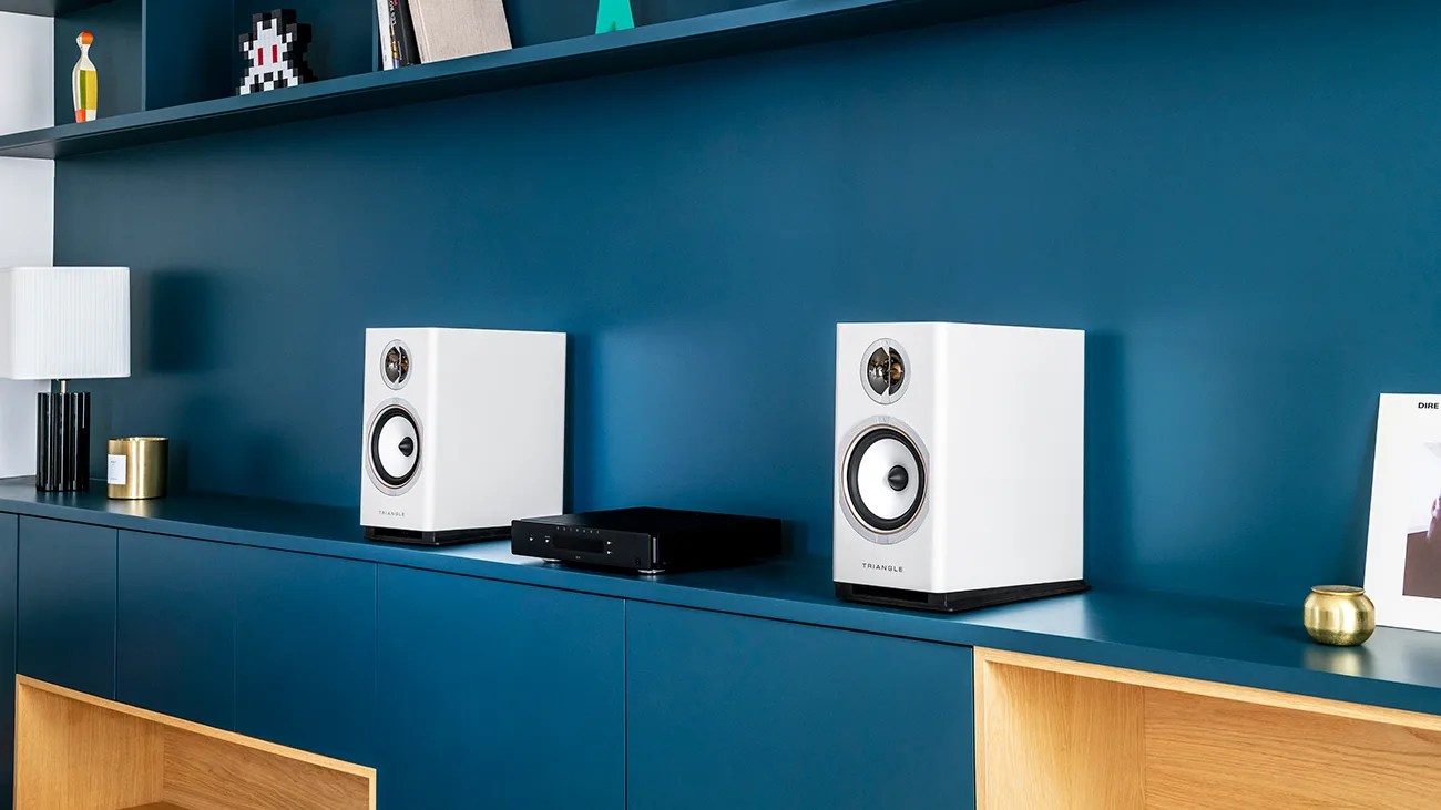 Pair of white Triangle bookshelf speakers on a blue cabinet with a black audio device between them.