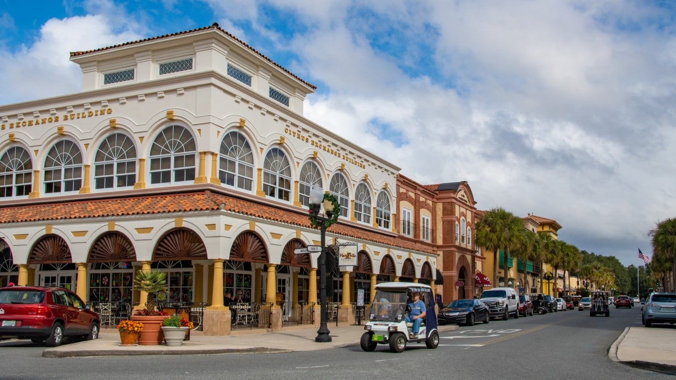 A bright street scene in a Florida town, featuring a prominent white building with arched windows and a red tile roof on the left, inscribed with 'CITRUS EXCHANGE BUILDING'. A red SUV is parked in front, and a white golf cart with two people drives down the street. The street is lined with palm trees and other Mediterranean-style buildings under a partly cloudy blue sky, with several cars and golf carts visible in the distance.