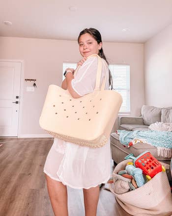 Person standing in a living room, holding a large tote bag. Dressed casually in sheer clothing, with children's toys visible in the background