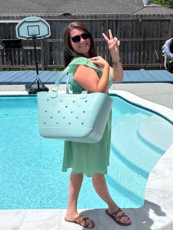 Woman by pool in a striped dress holding a large tote bag and making a peace sign