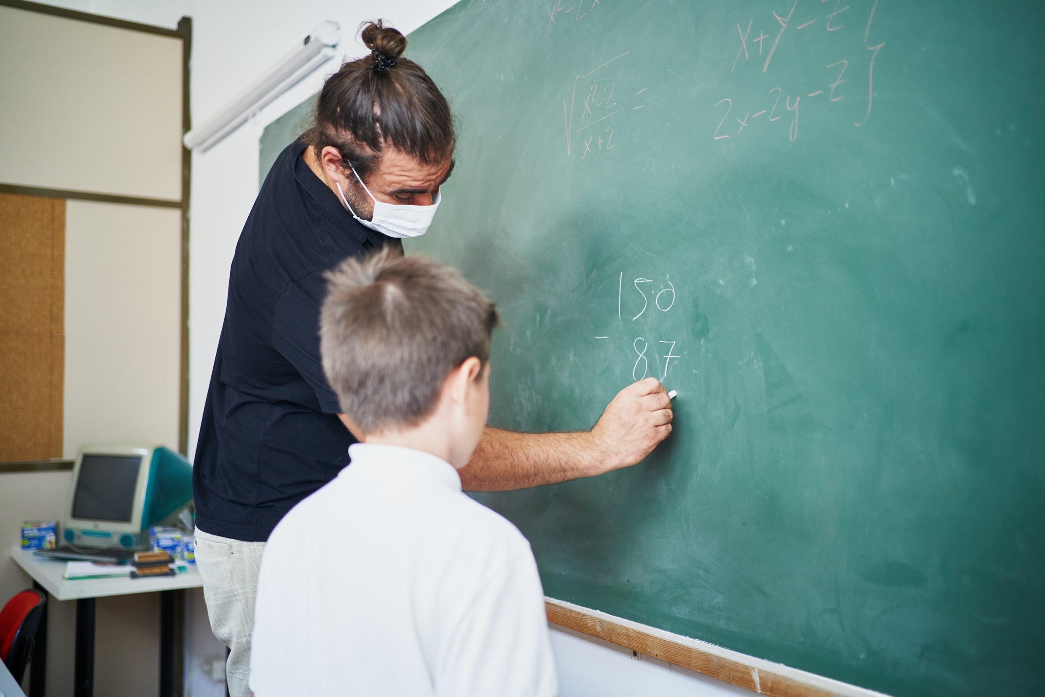 Teacher with a mask helps a young student solve math on a classroom chalkboard