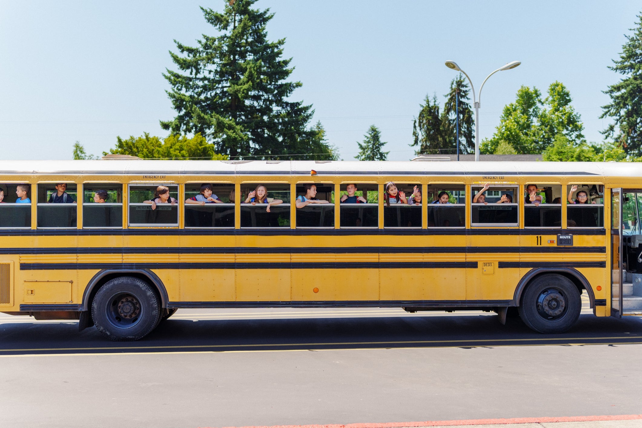 Children waving from the windows of a school bus parked on a street with trees in the background