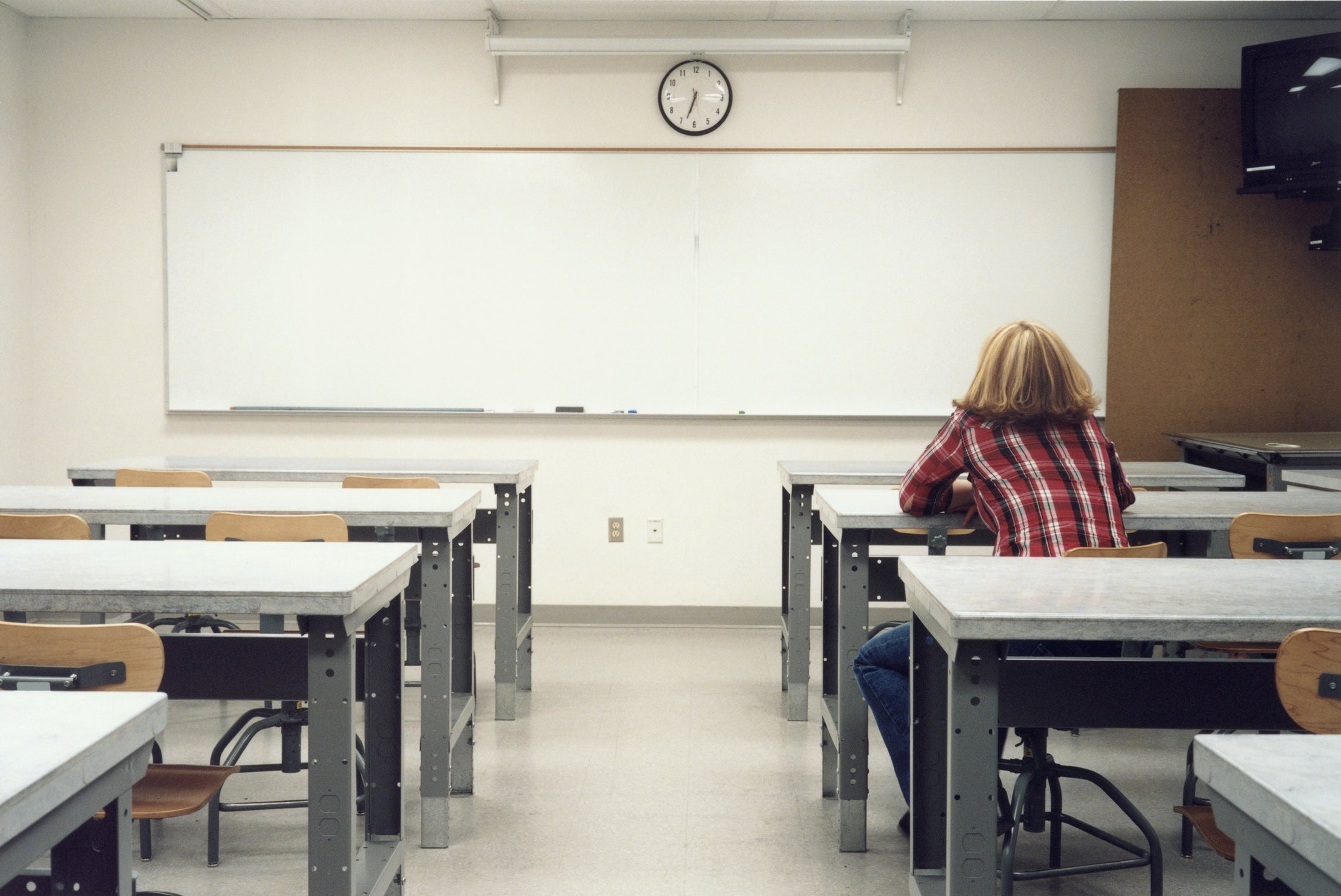 Person in a plaid shirt sits alone at a desk in a classroom facing a whiteboard and clock