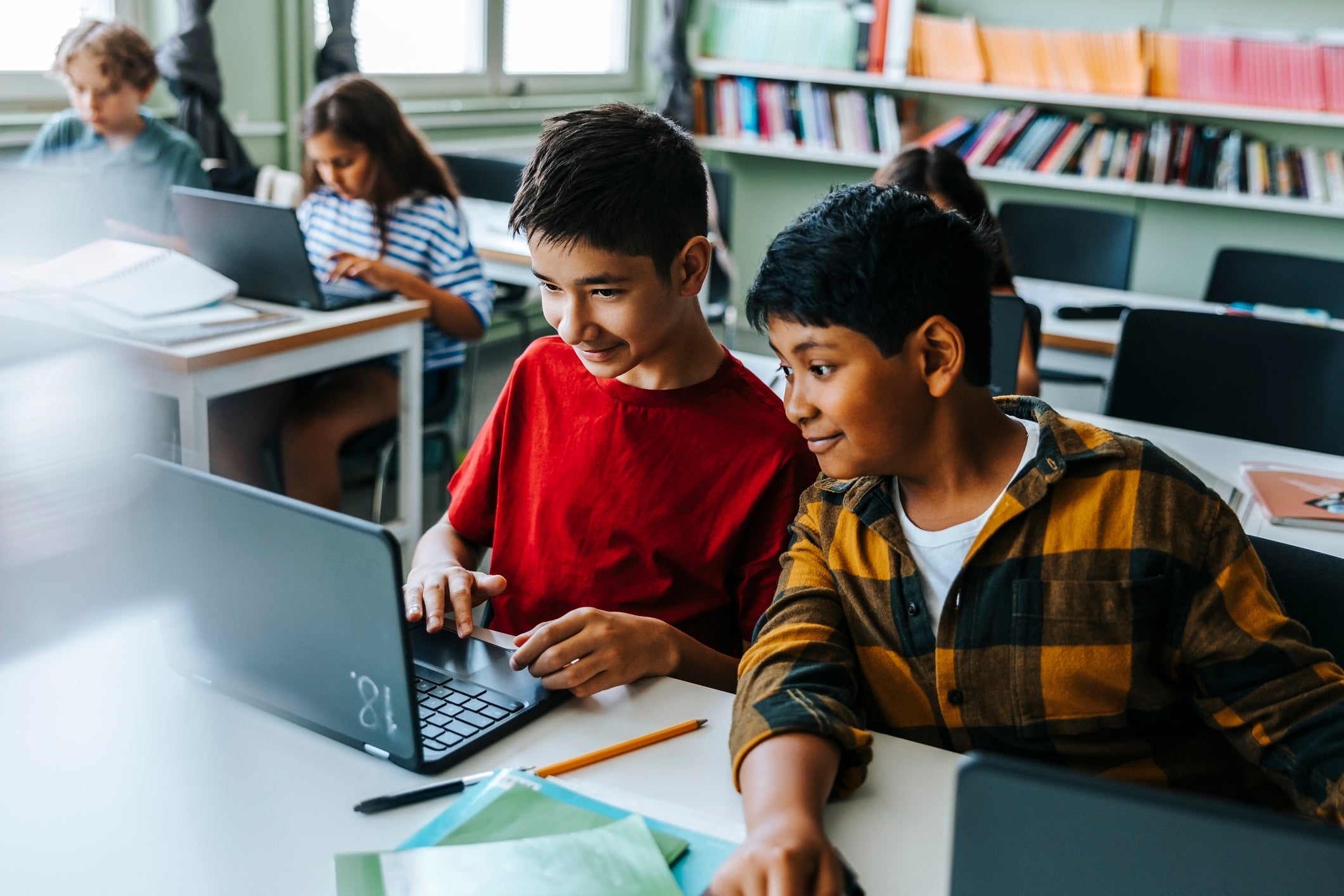 Two boys in a classroom sharing a laugh while looking at a laptop, surrounded by classmates also using computers