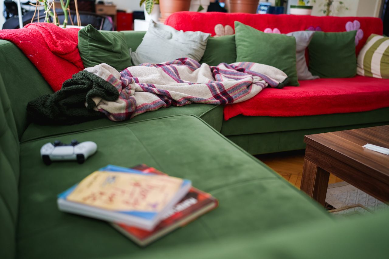 A living room setting with a green sectional sofa scattered cushions a blanket a game controller and books on a nearby table