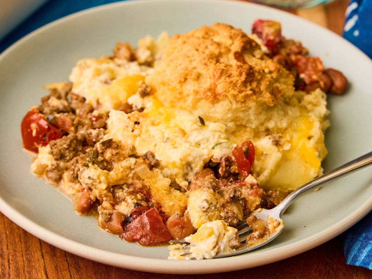 A plate with a serving of casserole containing meat vegetables and a biscuit topping with a fork resting on the plate