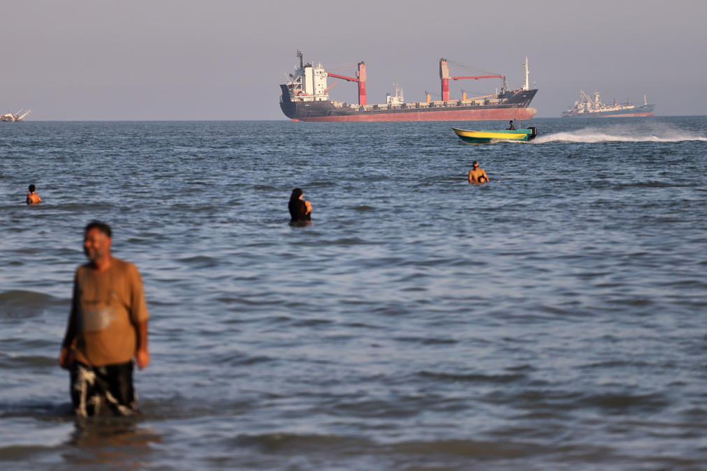 Residents swim in the waters of the Strait of Hormuz as a small motorboat passes cargo ships and other commercial vessels offshore near Bandar Abbas, Iran