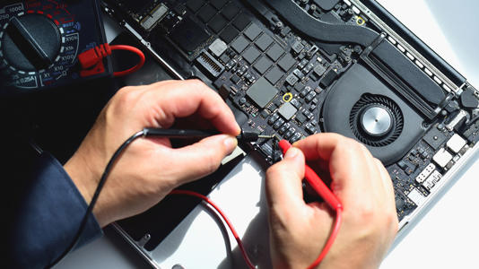 Technician testing an open laptop motherboard with a multimeter.