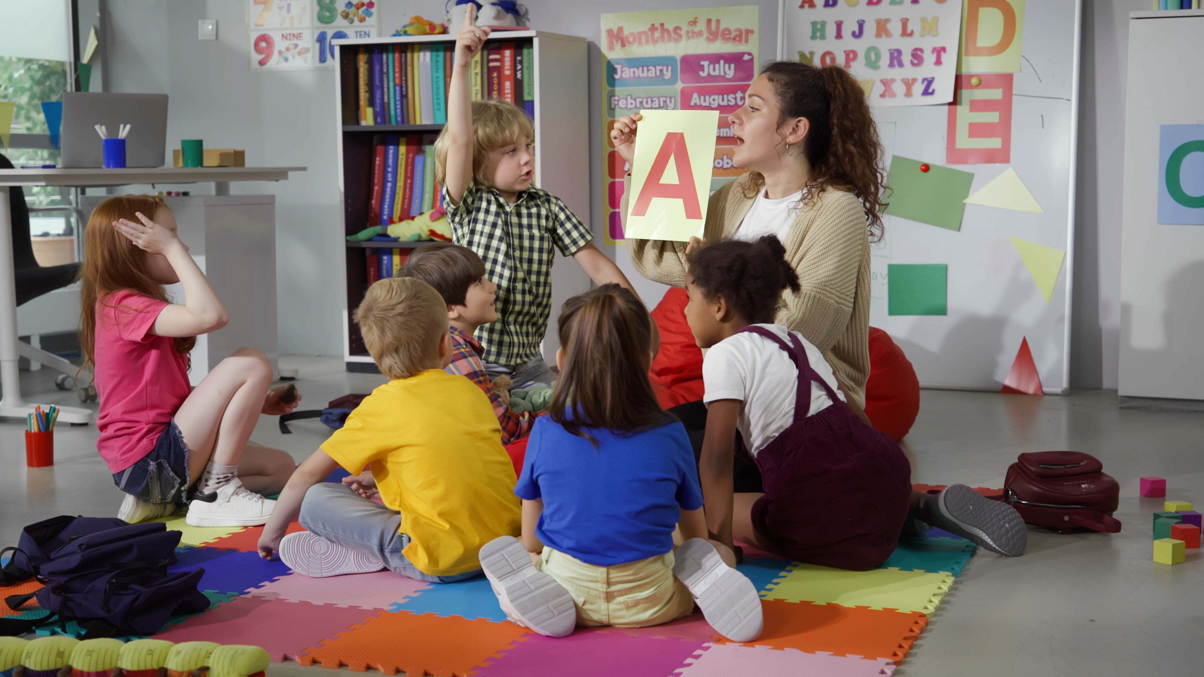 Teacher shows a large letter "A" to a group of young children sitting on a play mat in a classroom setting