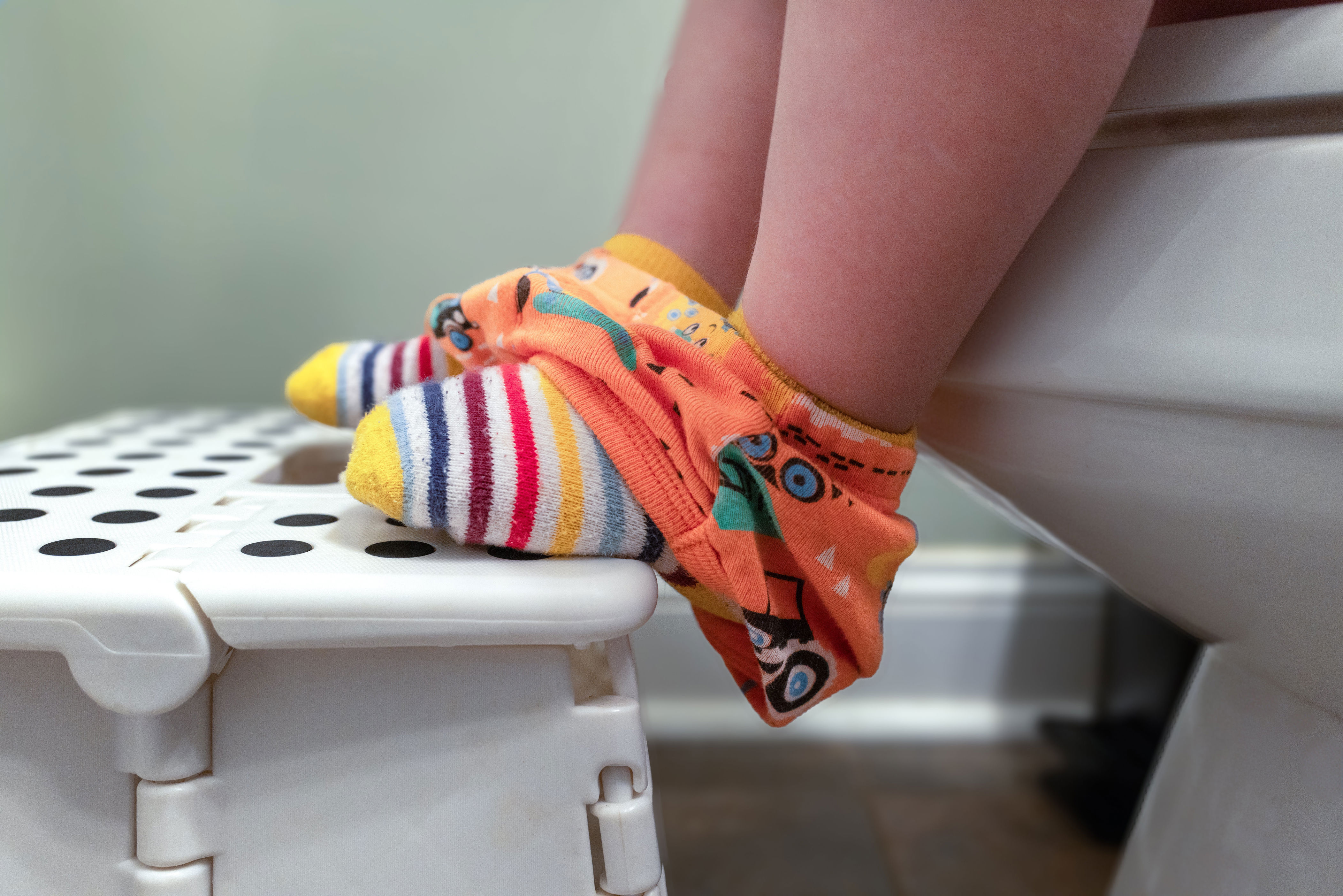 Child sitting on a toilet wearing striped socks and colorful pants, with feet on a step stool, suggesting potty training