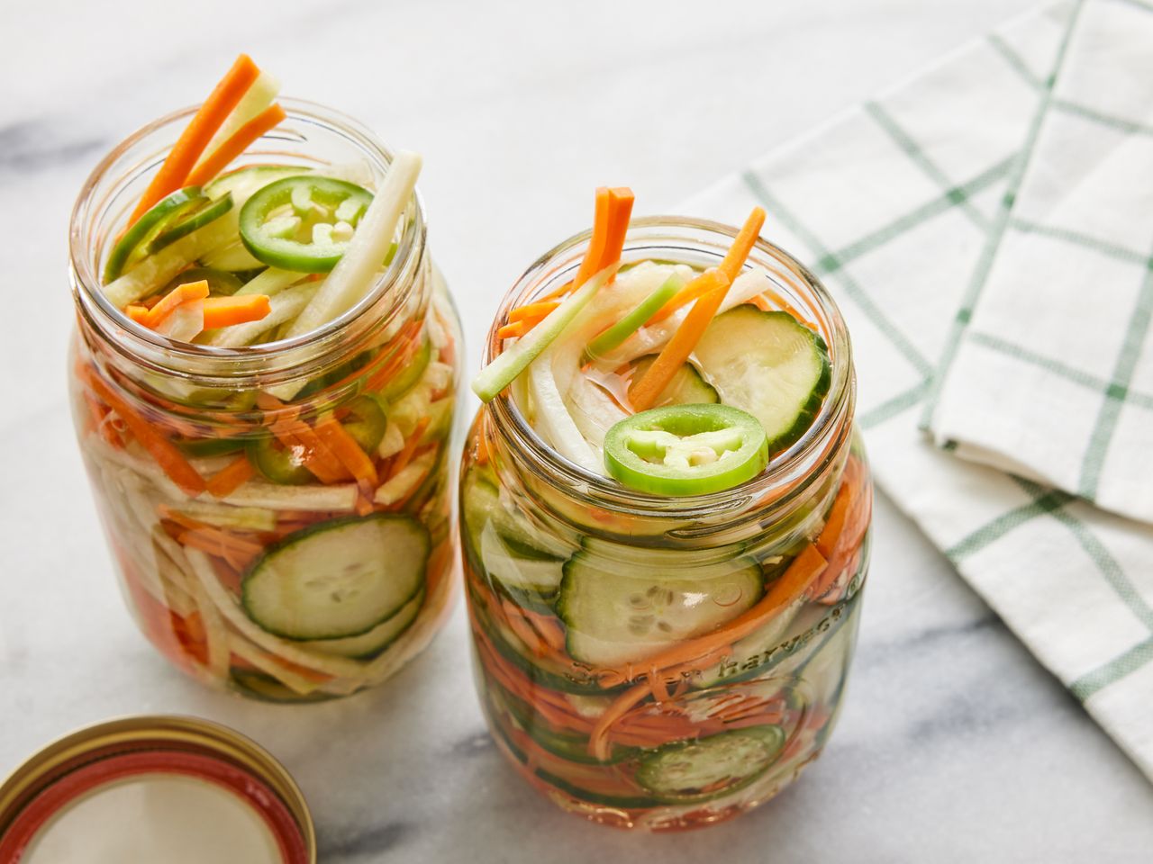Two glass jars filled with sliced vegetables in a pickling liquid placed on a countertop with a checkered cloth nearby