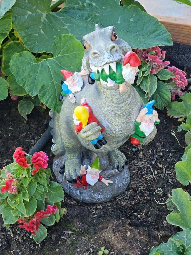 Garden sculpture of a dinosaur with colorful gnomes climbing on it, surrounded by lush plants and flowers