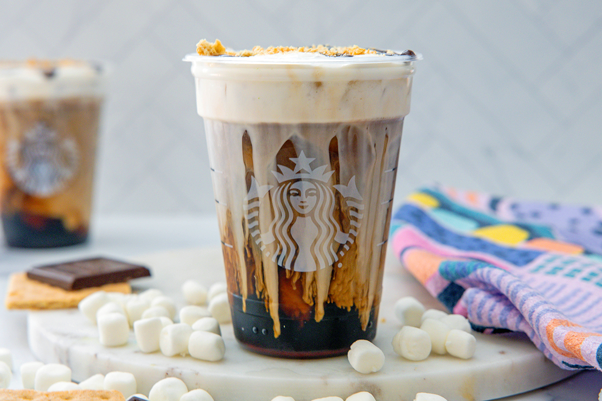 Landscape head-on view of a S'mores Cold Brew with second drink in background and mini marshmallows, graham crackers, and chocolate bars all around.