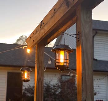 Hanging outdoor lanterns on a wooden pergola beam, illuminated at dusk, with a house and garden in the background. Ideal for patio decor