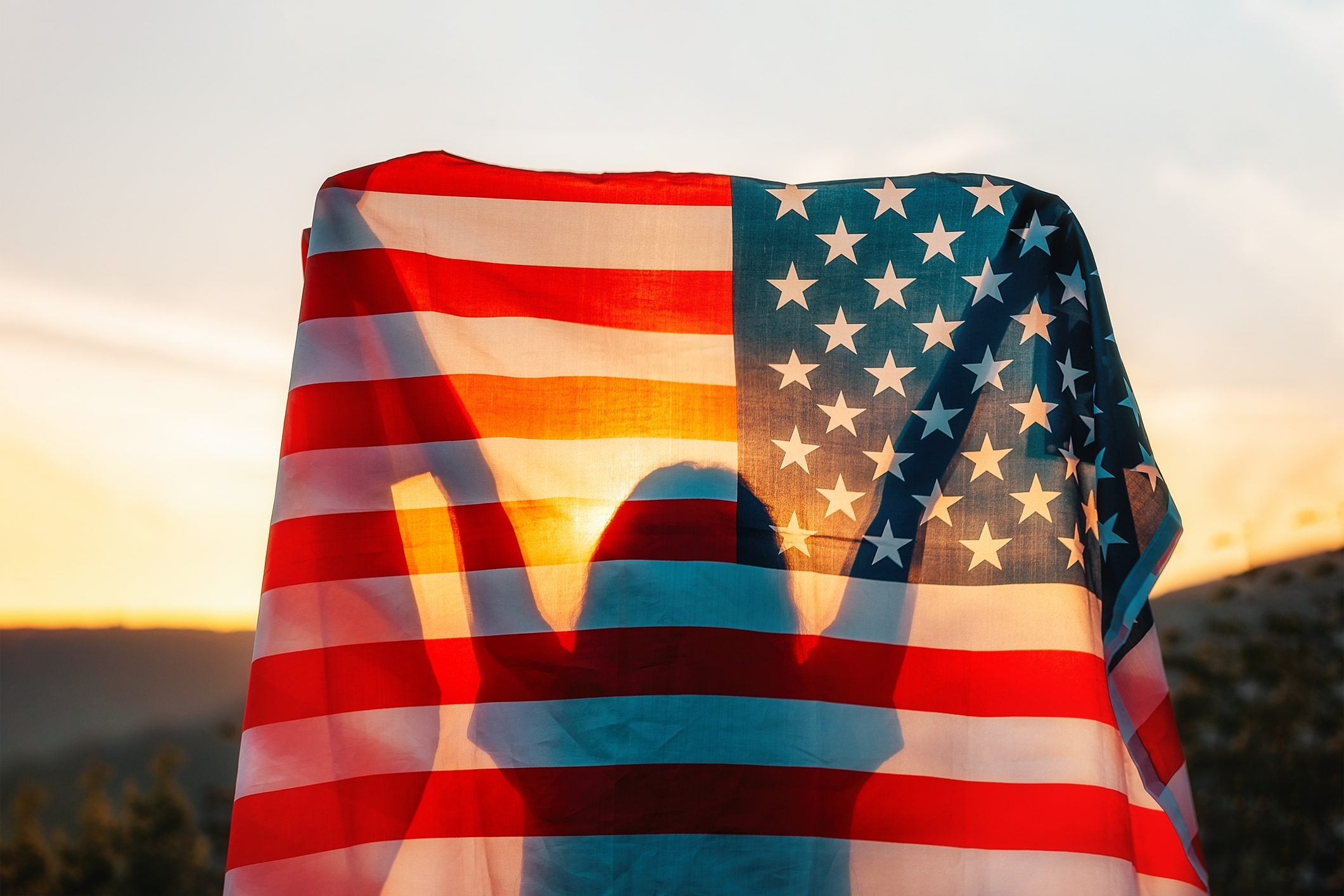 A person holds an American flag aloft against a sunset backdrop, creating a silhouette effect through the fabric.